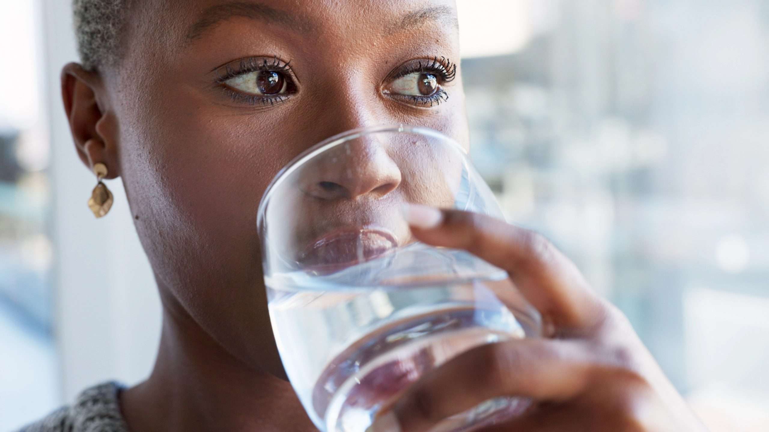 An adult woman drinking water in the foreground with a blurred background.